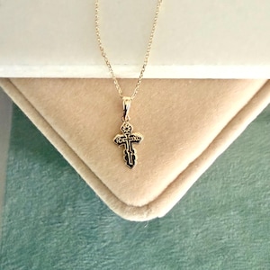May include: A gold-colored pendant necklace featuring a small, ornate cross. The cross has a detailed design with dark accents and is suspended from a delicate chain. The necklace is displayed on a light-colored, textured surface.