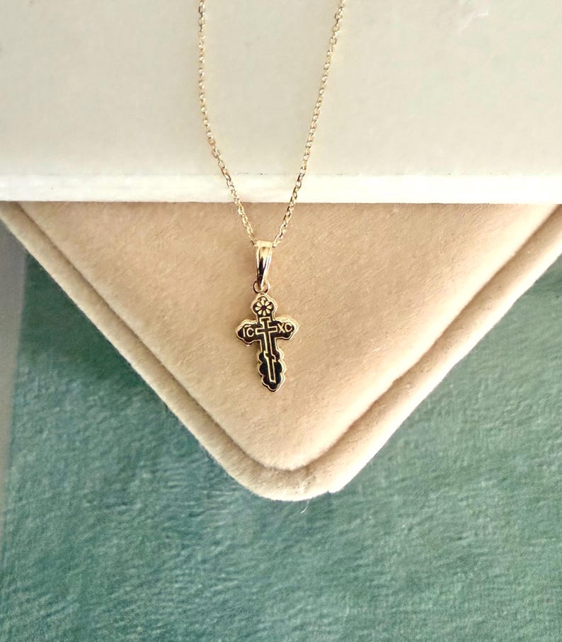 May include: A gold-colored pendant necklace featuring a small, ornate cross. The cross has a detailed design with dark accents and is suspended from a delicate chain. The necklace is displayed on a light-colored, textured surface.