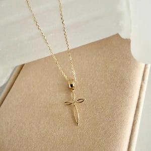 Puede incluir: Un delicado collar dorado con un colgante de cruz. El colgante tiene un dise&ntilde;o minimalista con l&iacute;neas curvas que forman la forma de la cruz. El collar se exhibe sobre una superficie de tonos neutros.