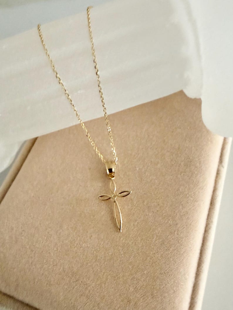 Puede incluir: Un delicado collar dorado con un colgante de cruz. El colgante tiene un dise&ntilde;o minimalista con l&iacute;neas curvas que forman la forma de la cruz. El collar se exhibe sobre una superficie de tonos neutros.