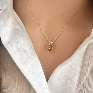 May include: A gold-colored necklace with a pendant featuring a light orange oval gemstone. The necklace is worn over a white collared shirt.