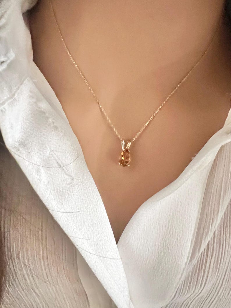 May include: A gold-colored necklace with a pendant featuring a light orange oval gemstone. The necklace is worn over a white collared shirt.