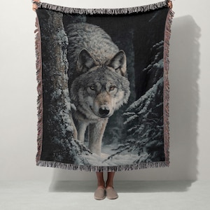 May include: A woven throw blanket featuring a detailed image of a gray wolf in a snowy forest. The wolf is the focal point, peering out from behind snow-covered trees. The blanket has a black background and a fringed edge.