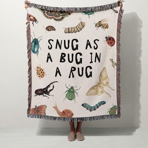 May include: A cream-colored throw blanket with a fringe edge, featuring various insect illustrations in shades of green, brown, and orange. The text "SNUG AS A BUG IN A RUG" is printed in black. The blanket is held up by a person.