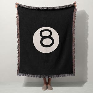 May include: Black throw blanket featuring a large white circle with the number 8 in the center. The blanket has a fringed edge and is held up by a person. The design resembles an 8-ball from a pool game.