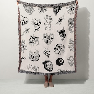 May include: Cream-colored throw blanket featuring a repeating pattern of black tattoo-style illustrations. Designs include a tiger, skull, rose, spiderweb, and an eight ball. The blanket has a fringed edge.