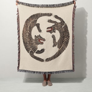 May include: A woven throw blanket in cream with a large circular graphic of two black tigers with striped patterns and red mouths. The blanket features a fringed edge with multi-colored threads along the top and bottom borders.