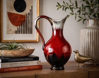 May include: A ceramic pitcher featuring a glossy finish that transitions from a deep black base to a vibrant red midsection, topped with a white drip glaze at the rim. The vessel has a curved handle and a flared spout, showcasing a smooth, reflective surface.