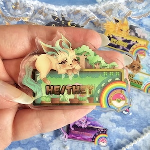 May include: A clear acrylic charm featuring a light yellow Eevee with green hair, sitting on a green and gold background with a rainbow and the text "HE/THEY".