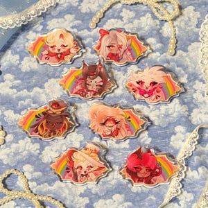 May include: A collection of nine clear acrylic pins featuring colorful cartoon characters with rainbow accents. Each pin has a unique character design with vibrant colors, set against a blue background with lace and pearl accents.