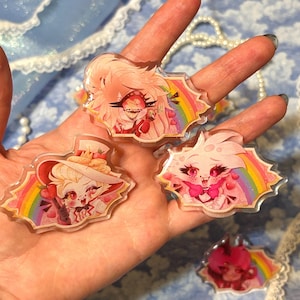 May include: Several clear acrylic charms featuring cartoon characters with pink and white color schemes, set against a rainbow background. The charms are held in a hand, showcasing their design and details. The charms have a cartoon theme.