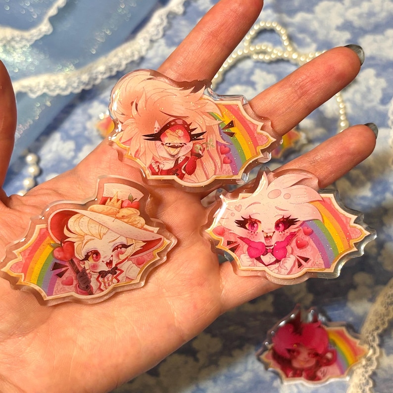 May include: Several clear acrylic charms featuring cartoon characters with pink and white color schemes, set against a rainbow background. The charms are held in a hand, showcasing their design and details. The charms have a cartoon theme.