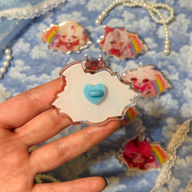 May include: A collection of colorful acrylic pins featuring anime-style characters with rainbow accents. The pins have a clear border and a light blue heart-shaped pin back. The background is a light blue floral pattern.
