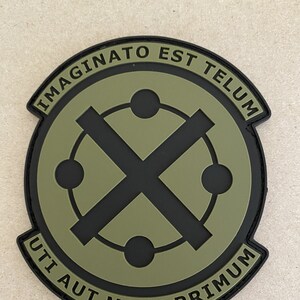 May include: A circular patch with a black border and a green interior. The patch features a black X intersecting a circle with four dots. The text "IMAGINATO EST TELUM" and "UTI AUT MORI PRIMUM" are arched around the top and bottom.