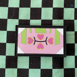 May include: A pink and green embroidered patch with a heart design. The patch has a black border and is made of fabric.