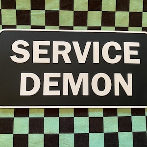 May include: Black and white rectangular sign with the text "SERVICE DEMON" in white raised letters.