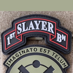 Puede incluir: Parche bordado negro y rojo con el texto "1st SLAYER BN" y un parche más pequeño debajo con el texto "IMAGINATO EST TELUM".