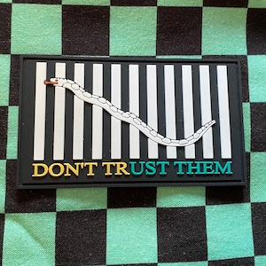 May include: Black and white striped patch with a white snake winding across it. The text "DON'T TRUST THEM" is written in yellow, blue, and green letters.