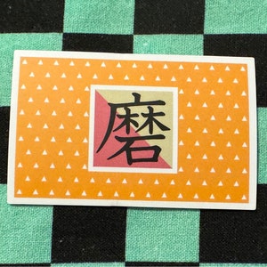 Peut inclure: Une carte rectangulaire avec un fond orange et un motif de triangles blancs. La carte a un carré rouge et blanc avec des caractères japonais noirs qui se lisent "Demon Slayer".