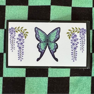 Peut inclure: Un autocollant blanc avec un papillon vert et des fleurs de glycine violettes sur un fond à carreaux noir et vert.
