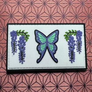 Puede incluir: Un parche blanco con borde negro presenta una mariposa morada con alas turquesa y dos racimos de flores de glicinia moradas a cada lado.