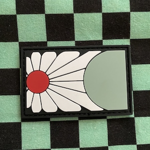 May include: A black and white patch with a white flower design and a red circle in the center. The flower has a green half-circle on the right side.