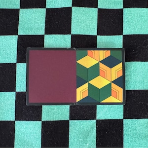 Peut inclure: Un autocollant avec un fond bordeaux et un motif géométrique de carrés verts, jaunes et orange. Le motif rappelle un design japonais traditionnel.