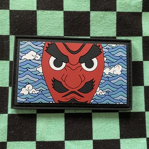 Puede incluir: Un parche rectangular con una máscara de demonio roja con detalles negros y ojos blancos, sobre un fondo de olas y nubes azules. El parche está enmarcado en un borde negro.