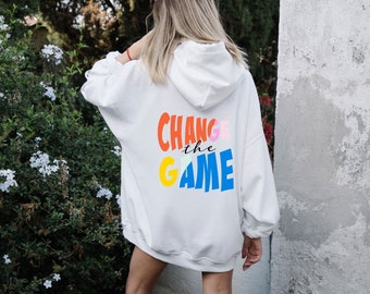 Könnte beinhalten: Weißer Kapuzenpullover mit dem Text "Change the Game" auf der Rückseite in bunten Buchstaben.