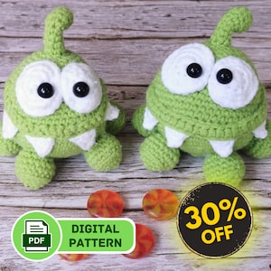 May include: Two green crocheted plush toys shaped like the Om Nom character from the game Cut the Rope. The toys have white eyes, white teeth, and a white antenna. The toys are sitting on a wooden surface with candy and a sign that says "Digital Pattern" and "30% Off".