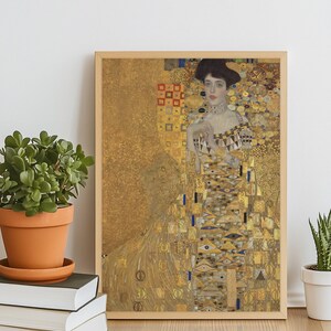May include: Framed print of a Gustav Klimt painting, featuring a woman in an ornate gold dress. The artwork has a gold background with geometric and organic patterns. The frame is light brown, and the print is displayed on a wooden surface.