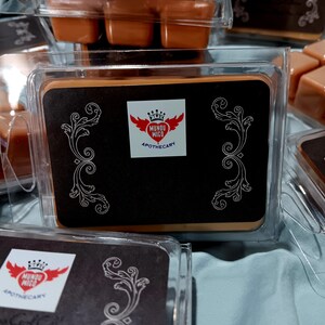 May include: A close-up of a brown wax melt with a black label that says "Mundo Migo Apothecary" with a red heart and crown logo. The wax melt is in a clear plastic container.
