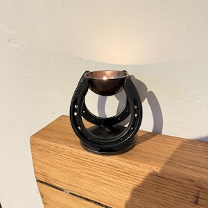 May include: A black metal candle holder with a horseshoe design. The holder has a copper-coloured bowl to hold the candle, which is lit. The candle holder is sitting on a wooden shelf.