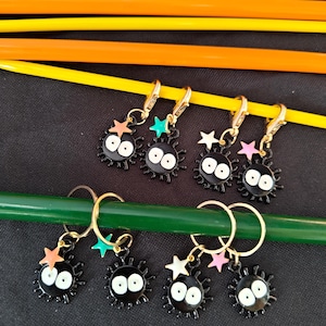May include: A collection of black and white charm stitch markers with gold-colored clasps, each featuring a cartoon-like character with star accents in various colors. The markers are arranged on a black surface with green, yellow, and orange knitting needles.