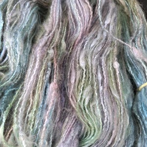 Puede incluir: Primer plano de una madeja de hilo teñido a mano en tonos azules, verdes, lavanda y blanco. El hilo tiene un aspecto suave y texturizado con sutiles variaciones de color y grosor, ideal para proyectos de tejido o ganchillo.