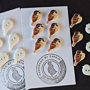 May include: A collection of decorative buttons in the shape of birds and bird silhouettes. The bird buttons are brown, beige, and white. The silhouette buttons are white. The cards have a logo that says "HANDMADE BY CATKIN MADE WITH LOVE."