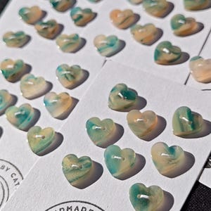 Pode incluir: Uma coleção de botões em forma de coração com uma mistura marmorizada de cores creme e azul-petróleo. Cada botão tem dois orifícios no centro e está preso a cartões de exibição brancos impressos com o texto HANDMADE BY CAT.