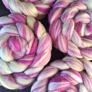Puede incluir: Tres trenzas circulares de lana cardada con una paleta de colores variados que incluye magenta vibrante, rosa pastel suave, blanco crema y verde lima pálido. La textura gruesa y esponjosa de la lana sin hilar es visible en todas las trenzas.