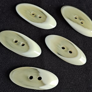 May include: Five oval buttons with two holes each. The buttons have a creamy white base colour with a swirl of light olive green. The buttons are arranged on a dark, textured surface.