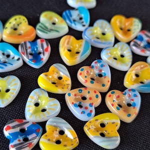 May include: A collection of heart-shaped buttons featuring various patterns in yellow, blue, orange, and white. Each button has two holes in the center and displays distinct designs including sunbursts, stripes, and colorful dots on a smooth surface.