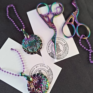 May include: Iridescent metal craft scissors and pendant necklaces with floral designs. The scissors and pendants have a rainbow finish. The pendants are attached to purple ball chains. The scissors and pendants are on white cards with a logo that says "HANDMADE BY CATKIN".