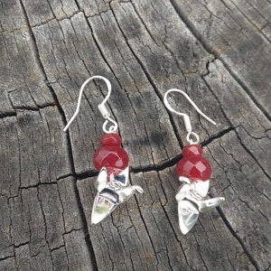 May include: A pair of silver earrings with red gemstone accents. The earrings feature a dangling design with a small, round gemstone at the top and a larger, faceted gemstone below.