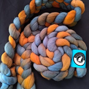 May include: Braided roving yarn in shades of blue, orange, and gray. The thick yarn is arranged in a large, looping braid. A small, light blue tag with a white cat logo and text is attached to the yarn.