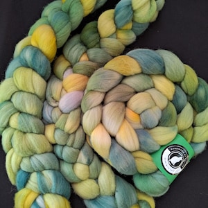 May include: Braided yarn in shades of yellow, green, and blue. The yarn is thick and appears to be hand-dyed. A green tag with white text and a logo is attached to the yarn. The yarn is displayed on a black surface.