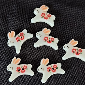 Pode incluir: Seis botões brancos em forma de coelhos a saltar. Cada botão tem dois orifícios, orelhas cor de pêssego e um pequeno padrão floral vermelho no corpo. Os coelhos têm detalhes simples de linhas pretas para os olhos e bigodes.