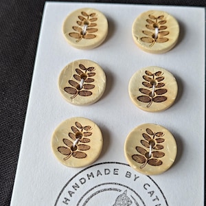 May include: Six round, light beige buttons arranged in two columns on a white card. Each button features a dark brown, laser-engraved fern leaf pattern and two central holes. The card displays a circular stamp with a cat silhouette and the text HANDMADE BY CATKIN.