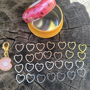 May include: A set of 30 heart-shaped stitch markers in gold, silver, and rose gold tones. The markers are arranged in rows on a wooden surface. A donut-shaped charm and a small tin container with a donut design are also included.