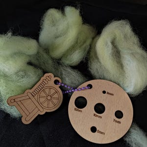 May include: Wooden yarn tools: a spindle charm and a circular gauge with holes labelled 5mm, 7mm, 8mm, 10mm, and 12mm. The tools are linked by a purple beaded chain. Light green wool fibers surround the tools, ideal for crafting.