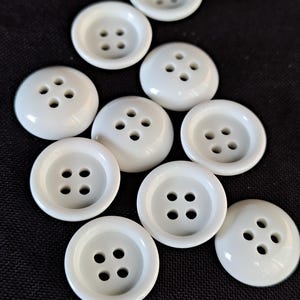 Op de afbeelding: Een verzameling van negen ronde, witte knopen, elk met een gladde, glanzende afwerking en vier gecentreerde gaten voor bevestiging. De knopen zijn verspreid gerangschikt en tonen hun uniforme cirkelvormige vorm en verhoogde randen.