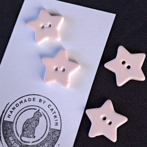 May include: Four light pink star-shaped buttons with two holes each. Two buttons are attached to a white rectangular card featuring a circular black stamp with a cat illustration and the text HANDMADE BY CATKIN MADE WITH LOVE.